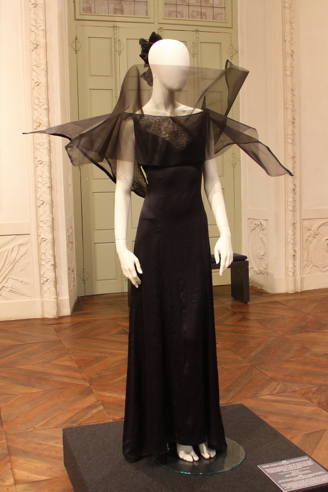 La_ Rai_Racconta_L'Italia_05.JPG - MINA   Abito lungo in satin nero, corpino formato da due strati di crine nero con punte modellabili. Acconciatura a grappolo di fiori in organza e tulle nero con nastro raso nero.   Questo look fa parte della serie di abiti, entrati nella storia del costume, ideati da Piero Gherardi, premio Oscar per "La dolce vita" e "Otto e mezzo", per la serie più famosa di Caroselli Barilla.   I filmati vennero girati nelle location che il regista e scenografo aveva identificato per "Il viaggio di G. Mastorna, film mai realizzato di Federico Fellini. II restauro dei capi è stato eseguito da Gabriele Mayer, all'epoca assistente di Gherardi. Programma Carosello Barilla "Ta-ra-ta-tà (fumo blu)" Anno 1967 Sartoria G.P. 11 Archivio Collezione Sartoria The One Srl Costumi, scenografia e regia Piero Gherardi